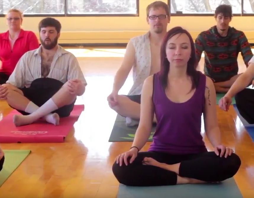 A group of college people meditate on yoga matts in brightly lit wood floored room