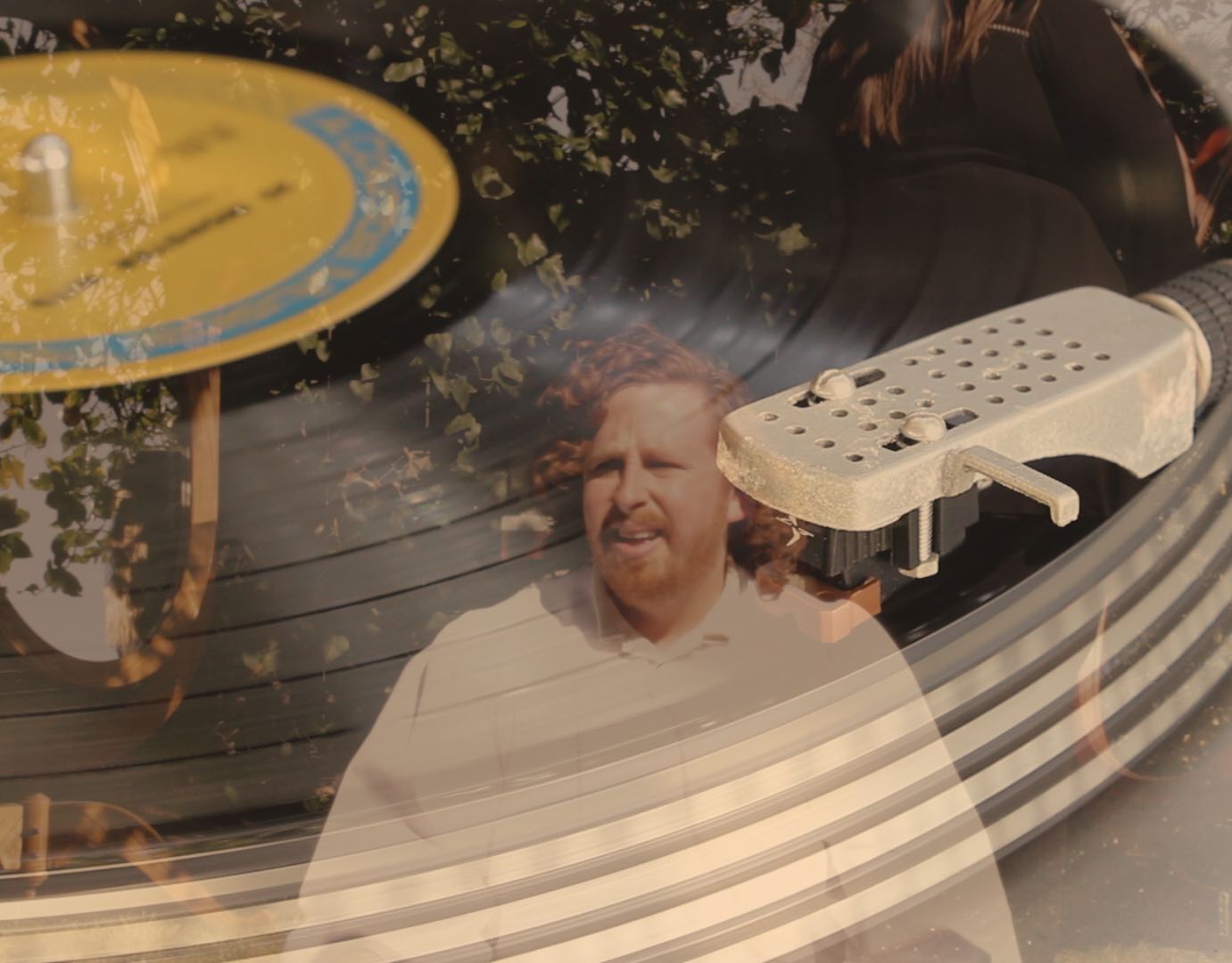 Still of a bewildered man sitting up in a white robe with a partially opaque record player over the image